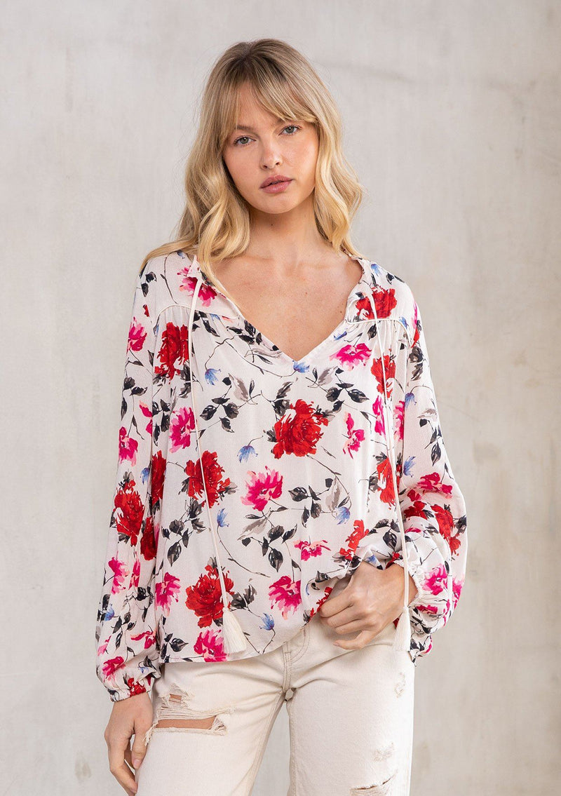 [Color: Cream/Red] A model wearing a classic peasant blouse in a romantic red rose floral print. With long sleeves, a ruffled neckline with tassel ties, and a flowy, relaxed fit. 