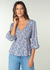 [Color: Blue/Coral] A front facing image of a brunette model wearing a classic bohemian peplum blouse in a blue and coral floral print. With long three quarter length sleeves, a v neckline, and a button front. 