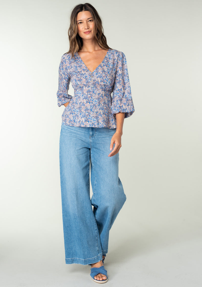 [Color: Blue/Coral] A full body front facing image of a brunette model wearing a classic bohemian peplum blouse in a blue and coral floral print. With long three quarter length sleeves, a v neckline, and a button front. 