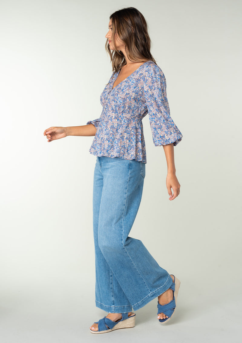 [Color: Blue/Coral] A side facing image of a brunette model wearing a classic bohemian peplum blouse in a blue and coral floral print. With long three quarter length sleeves, a v neckline, and a button front. 
