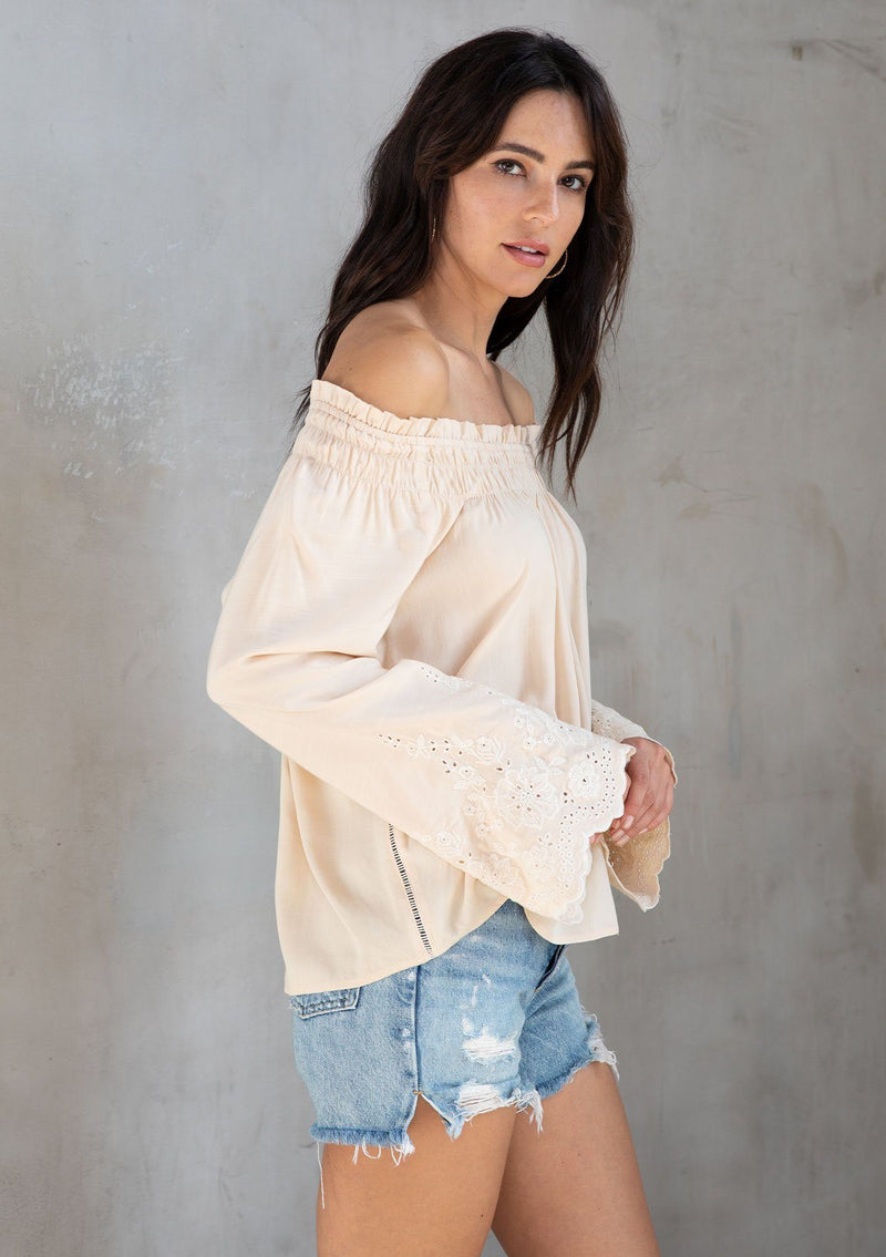 [Color: Natural] A model wearing a classic bohemian natural top with a smocked off the shoulder neckline and long embroidered bell sleeves. 