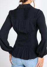 [Color: Black] A model wearing a black button front shirt with long sleeves and a waist cinching smocked detail at the front and back waist. 