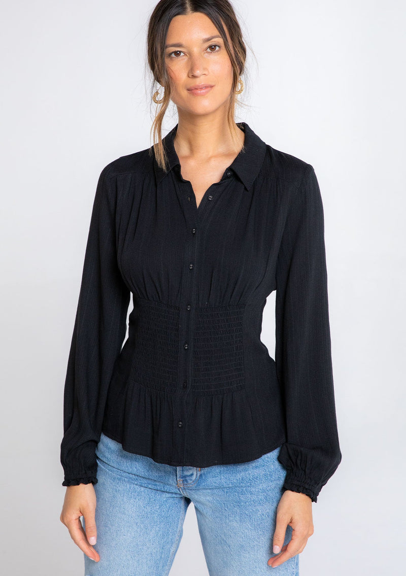 [Color: Black] A model wearing a black button front shirt with long sleeves and a waist cinching smocked detail at the front and back waist. 