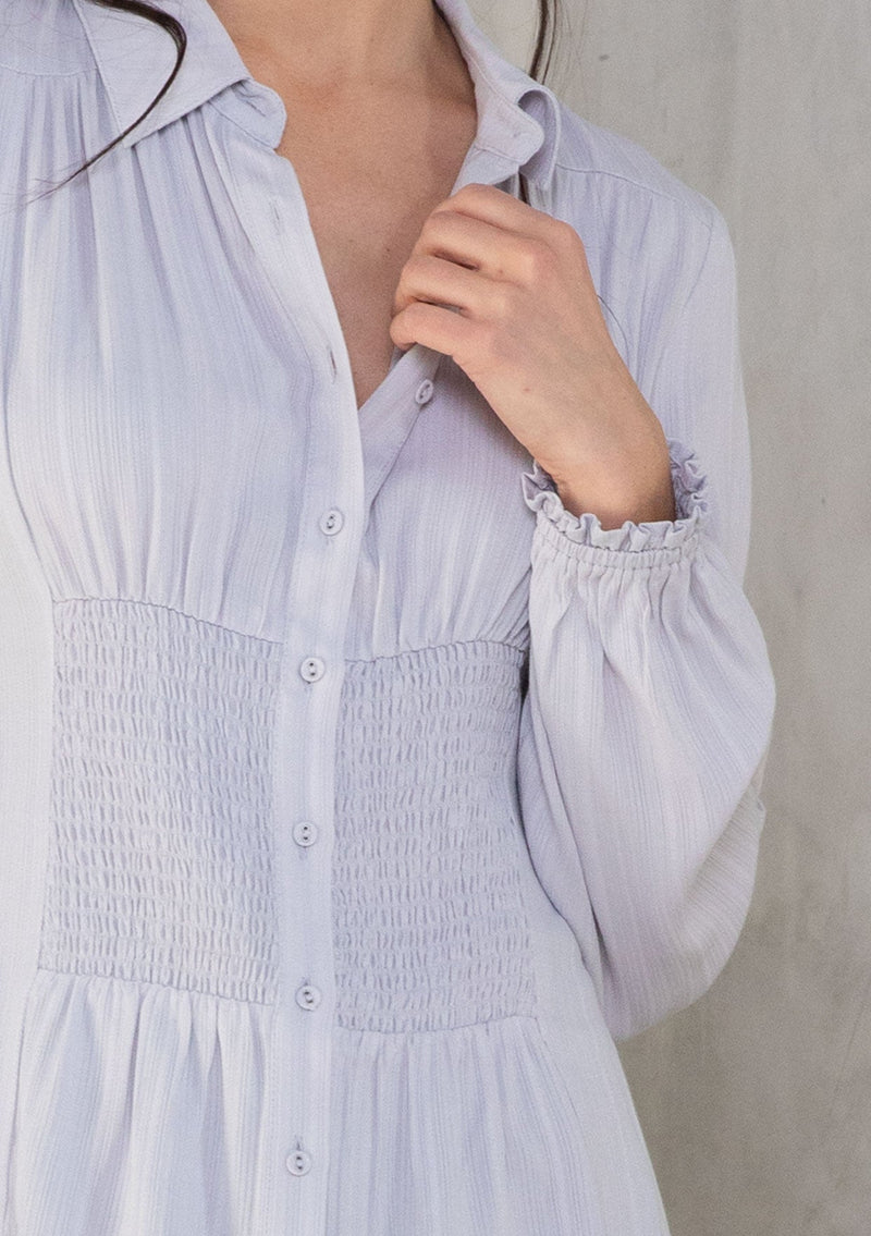 [Color: Dusty Lilac] A model wearing a light purple button front shirt with long sleeves and a waist cinching smocked detail at the front and back waist. 