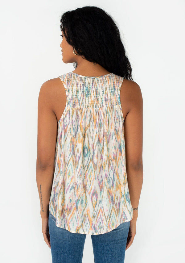 [Color: Natural/Teal] A back facing image of a brunette model wearing a casual resort tank top in a multi color bohemian diamond print. With a v neckline and a smocked yoke detail. 