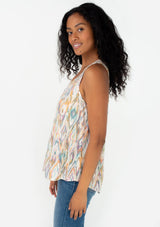 [Color: Natural/Teal] A side facing image of a brunette model wearing a casual resort tank top in a multi color bohemian diamond print. With a v neckline and a smocked yoke detail. 