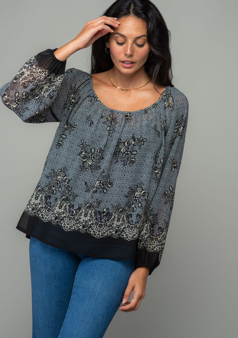[Color: Blue/Black] A front facing image of a brunette model wearing a chiffon bohemian blouse in a black and blue floral border print. With long sleeves and a wide elastic neckline. 