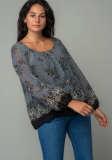 [Color: Blue/Black] A half body front facing image of a brunette model wearing a chiffon bohemian blouse in a black and blue floral border print. With long sleeves and a wide elastic neckline. 