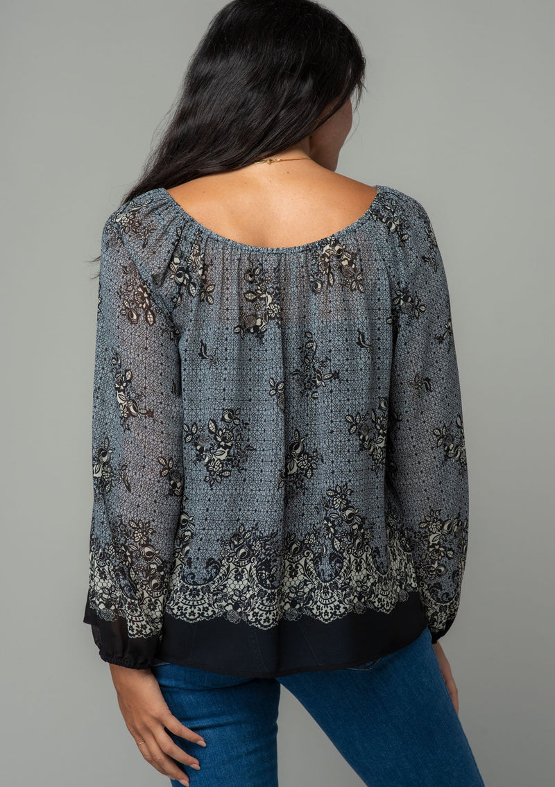 [Color: Blue/Black] A back facing image of a brunette model wearing a chiffon bohemian blouse in a black and blue floral border print. With long sleeves and a wide elastic neckline. 