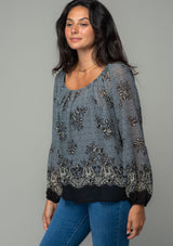 [Color: Blue/Black] A side facing image of a brunette model wearing a chiffon bohemian blouse in a black and blue floral border print. With long sleeves and a wide elastic neckline. 