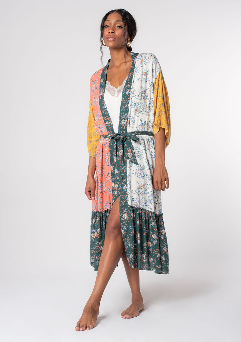 [Color: Coral/Off White] A model wearing a coral, white, yellow and green floral color block print bohemian kimono lounge robe with a waist belt and ruffled hemline.