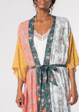 [Color: Coral/Off White] A model wearing a coral, white, yellow and green floral color block print bohemian kimono lounge robe with a waist belt and ruffled hemline.