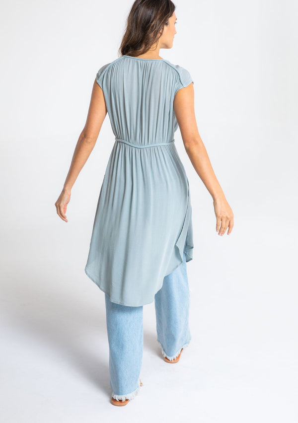 [Color: Blue Fog] A model wearing a floaty blue bohemian sheer cap sleeve tunic top with button front and braided trim. 