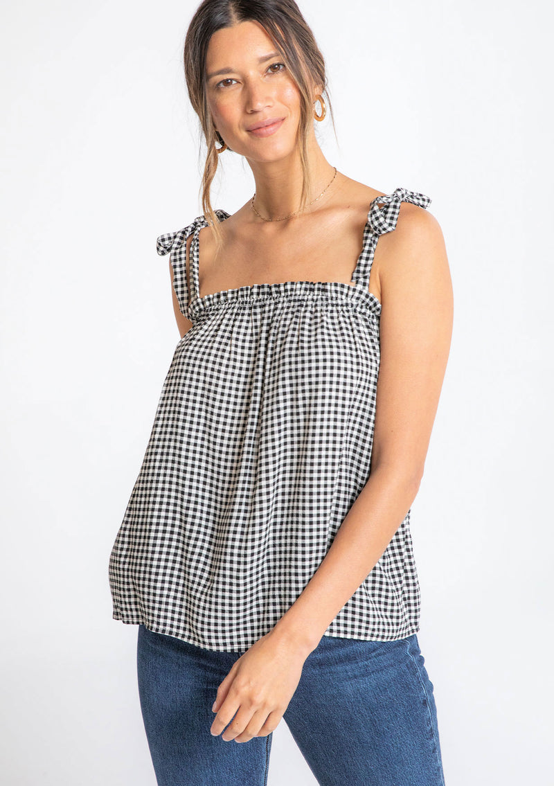 [Color: Black/Natural] A model wearing a black and white small checkered gingham print tank top with a tie shoulder detail. 