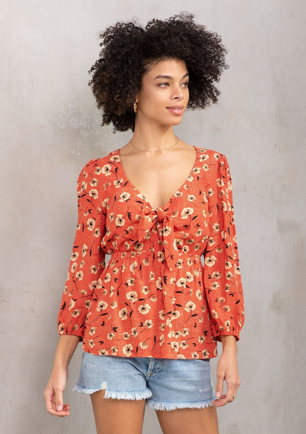 [Color: Red/Natural] A model wearing a vintage inspired blouse in a red floral print. With a sweetheart neckline, a tie front detail, and long sleeves. 