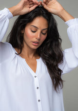 [Color: Chalk] A close up front facing image of a brunette model wearing a soft and silky white button front blouse with long sleeves, smocked wrist cuffs, and a split v neckline. 