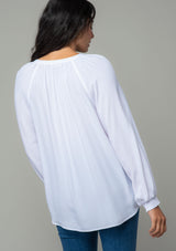 [Color: Chalk] A back facing image of a brunette model wearing a soft and silky white button front blouse with long sleeves, smocked wrist cuffs, and a split v neckline. 
