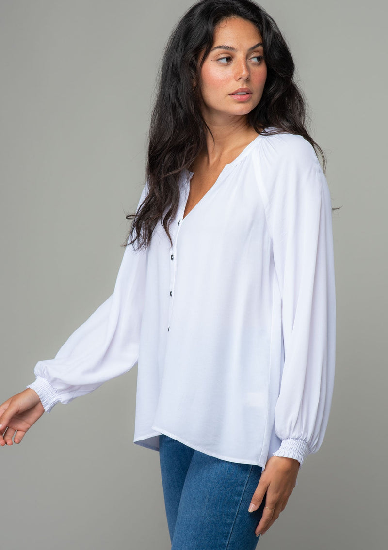 [Color: Chalk] A side facing image of a brunette model wearing a soft and silky white button front blouse with long sleeves, smocked wrist cuffs, and a split v neckline. 