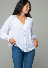 [Color: Chalk] A front facing image of a brunette model wearing a soft and silky white button front blouse with long sleeves, smocked wrist cuffs, and a split v neckline. 