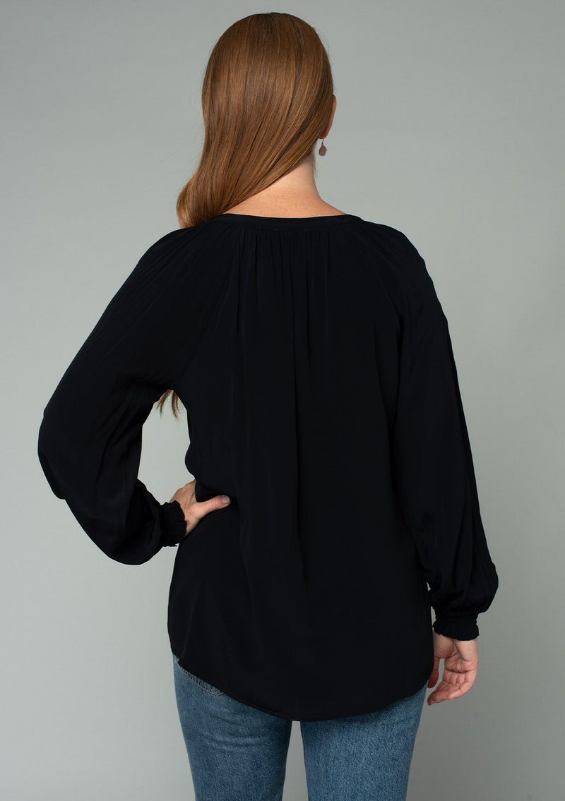 [Color: Black] A back facing image of a red headed model wearing a soft and silky black button front blouse with long sleeves, smocked wrist cuffs, and a split v neckline.