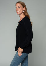 [Color: Black] A side facing image of a red headed model wearing a soft and silky black button front blouse with long sleeves, smocked wrist cuffs, and a split v neckline.