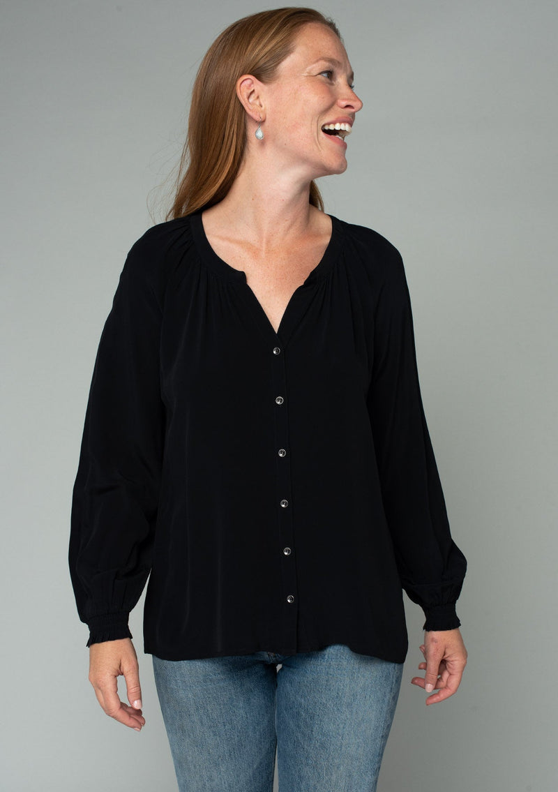 [Color: Black] A half body front facing image of a red headed model wearing a soft and silky black button front blouse with long sleeves, smocked wrist cuffs, and a split v neckline.