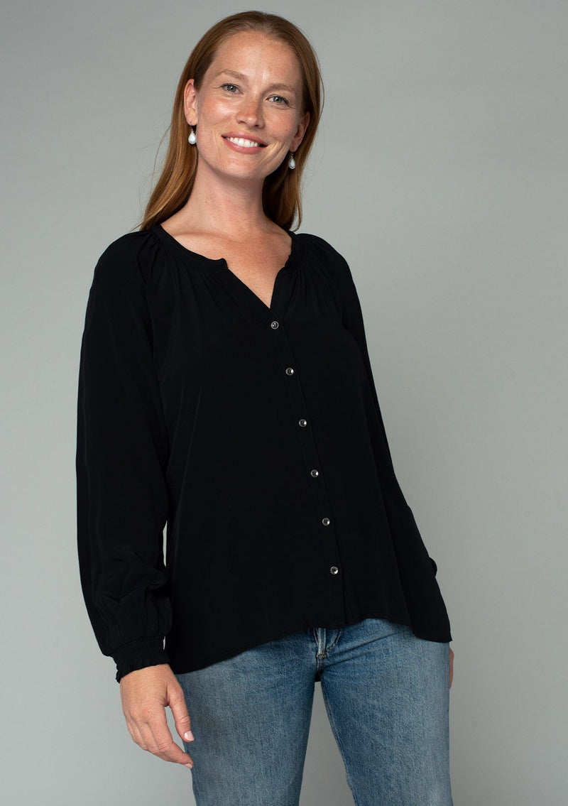[Color: Black] A front facing image of a red headed model wearing a soft and silky black button front blouse with long sleeves, smocked wrist cuffs, and a split v neckline.