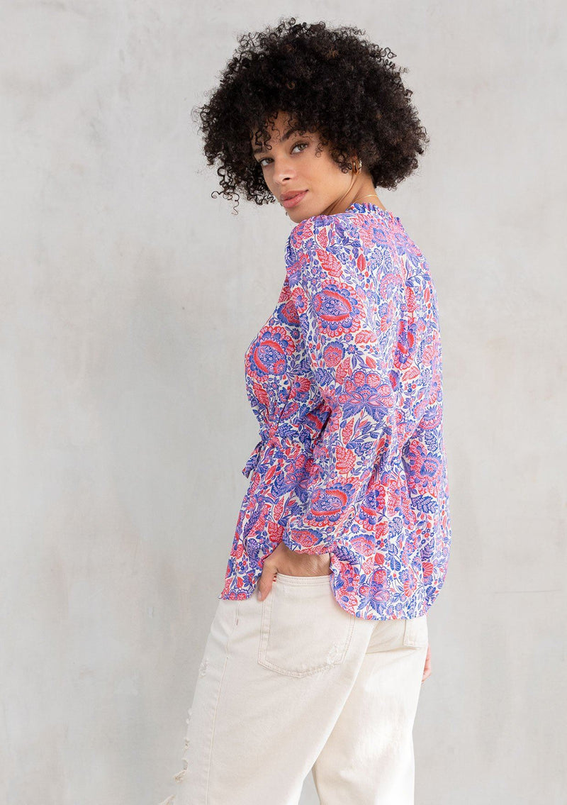 [Color: Ivory/Cobalt] A model wearing a flirty bohemian top in a red and blue paisley print. With a ruffle trimmed v neckline and a drawstring tie waist. 