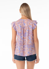 [Color: Ivory/Coral] A back facing image of a blonde model wearing a bohemian spring top in a retro inspired purple floral print. With short double flutter cap sleeves, a split v neckline with tassel ties, and a relaxed fit. 