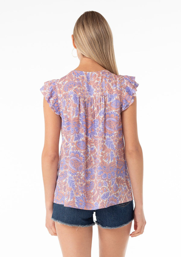 [Color: Ivory/Coral] A back facing image of a blonde model wearing a bohemian spring top in a retro inspired purple floral print. With short double flutter cap sleeves, a split v neckline with tassel ties, and a relaxed fit. 