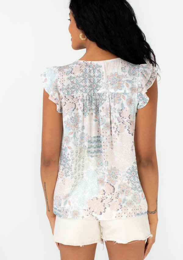 [Color: Dusty Blue/Teal] A back facing image of a brunette model wearing a bohemian spring blouse in a pastel blue and teal floral print. With short flutter sleeves, a split v neckline with tassel ties, and a relaxed fit. 