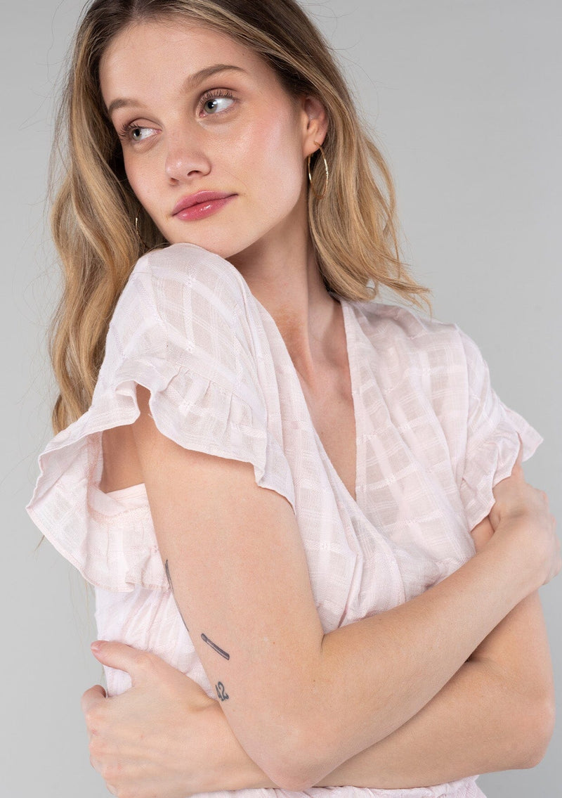[Color: Light Pink] A close up side facing image of a blonde model wearing a light pink cotton bohemian top with short ruffled sleeves, a smocked elastic waist, and a surplice faux wrap front with a v neckline.
