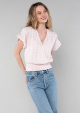 [Color: Light Pink] A half body front facing image of a blonde model wearing a light pink cotton bohemian top with short ruffled sleeves, a smocked elastic waist, and a surplice faux wrap front with a v neckline.