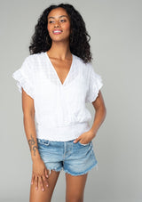 [Color: White] A front facing image of a brunette model wearing a bohemian white cotton top with short ruffled sleeves and a faux wrap front.