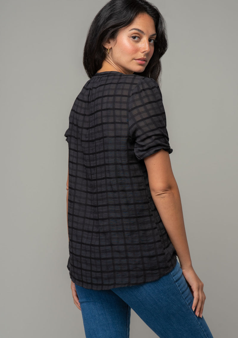 [Color: Black] A back facing image of a brunette model wearing a bohemian black cotton button front top with short puff sleeves. 