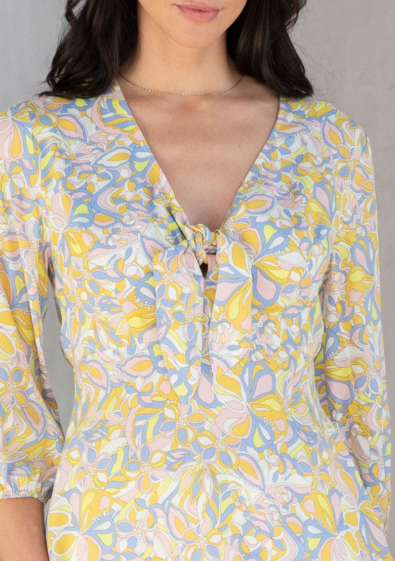 [Color: Blue/Lemon] A model wearing a blue and yellow retro floral print peplum top with a tie front detail. 