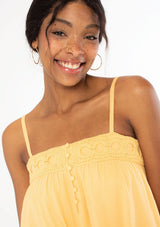 [Color: Sherbert] A woman wearing a yellow flowy bohemian tank top with crochet trim, gold toned hardware, and adjustable spaghetti straps.