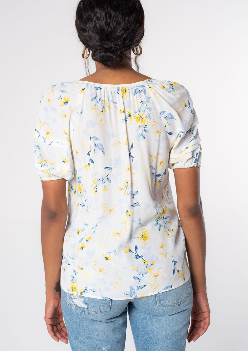 [Color: Cream/Yellow] A model wearing a white, yellow, and blue floral print puff sleeve blouse with tassel ties. 