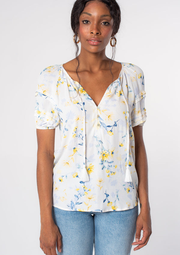 [Color: Cream/Yellow] A model wearing a white, yellow, and blue floral print puff sleeve blouse with tassel ties. 