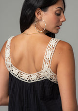 [Color: Black/Natural] A close up back facing image of a brunette model wearing a black flowy cotton gauze tank top with natural crochet trim and crochet tank top straps.