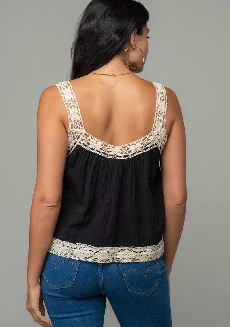 [Color: Black/Natural] A back facing image of a brunette model wearing a black flowy cotton gauze tank top with natural crochet trim and crochet tank top straps.