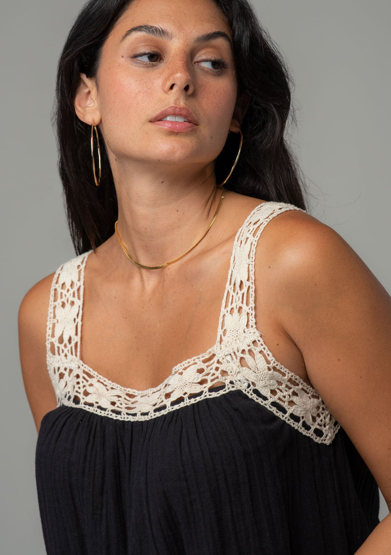[Color: Black/Natural] A close up front facing image of a brunette model wearing a black flowy cotton gauze tank top with natural crochet trim and crochet tank top straps.