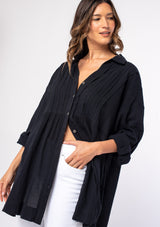 [Color: Black] A model wearing a relaxed black cotton tunic shirt with a button front, long rolled sleeves, and pleated details.