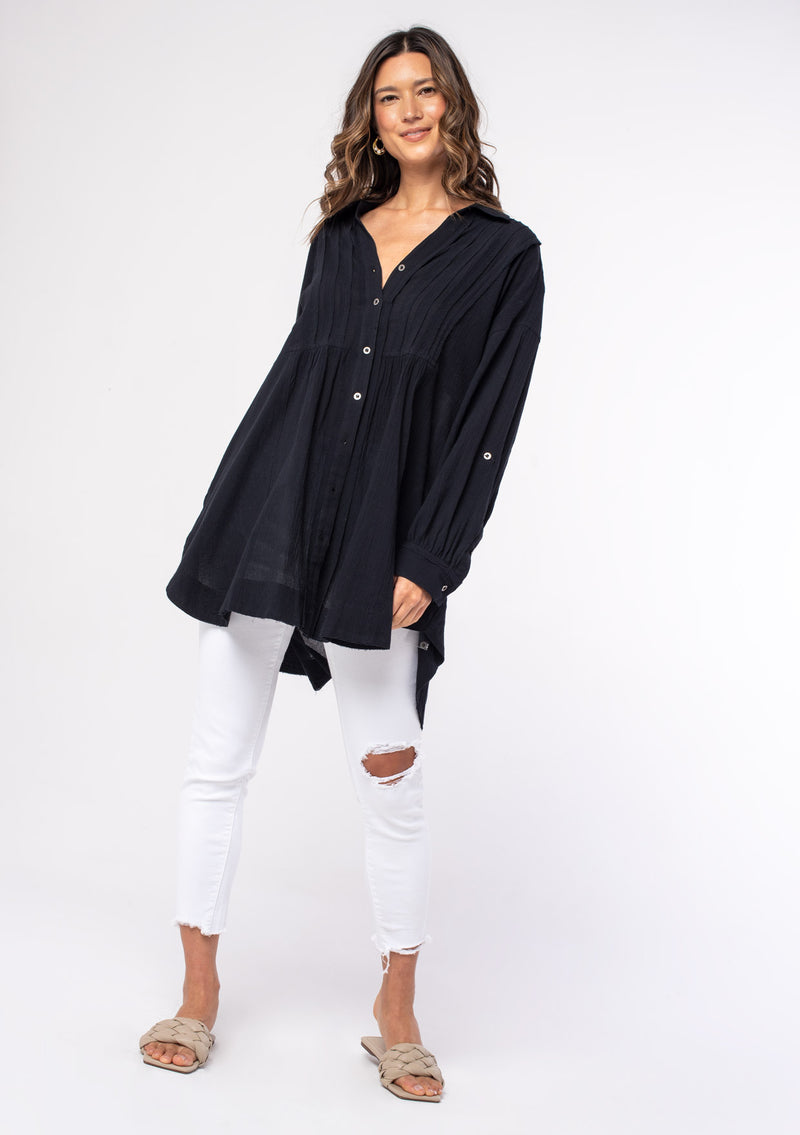 [Color: Black] A model wearing a relaxed black cotton tunic shirt with a button front, long rolled sleeves, and pleated details.