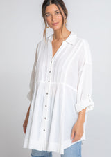 [Color: Off White] A model wearing a relaxed cotton tunic shirt with a button front, long rolled sleeves, and pleated details. 