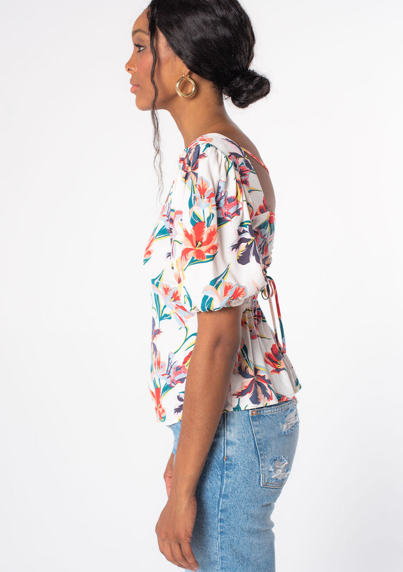 [Color: Ivory/Red] A woman wearing a white bohemian top with red floral print. With short puff sleeves and an open back detail with adjustable ties.
