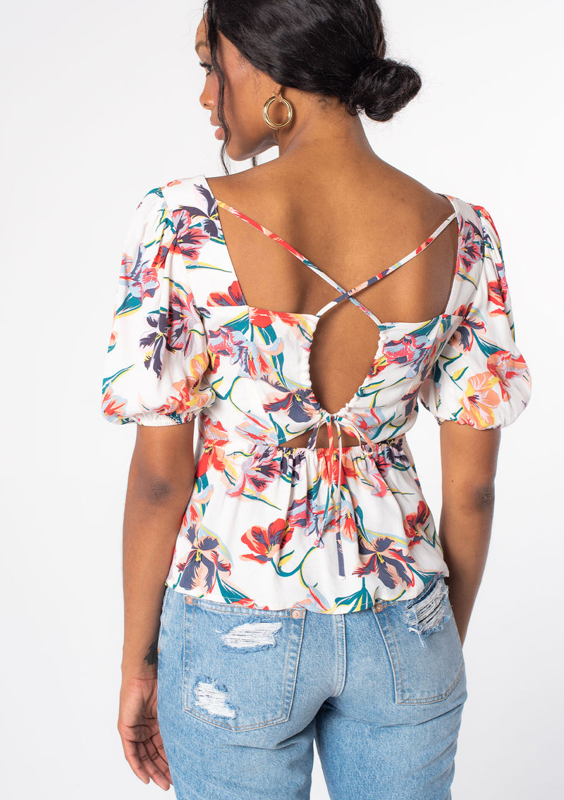 [Color: Ivory/Red] A woman wearing a white bohemian top with red floral print. With short puff sleeves and an open back detail with adjustable ties.