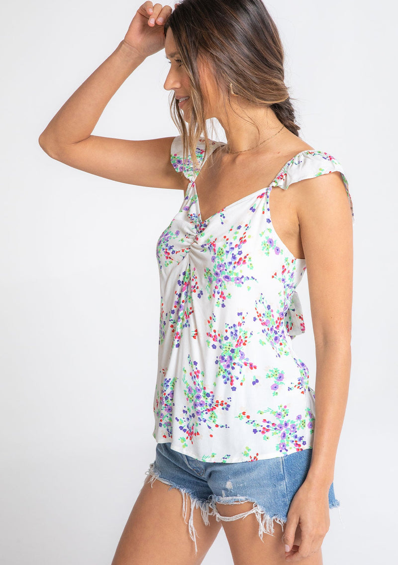 [Color: Ivory/Violet] A model wearing a white bohemian short flutter sleeve top with a purple floral print throughout and an adjustable back tie. 