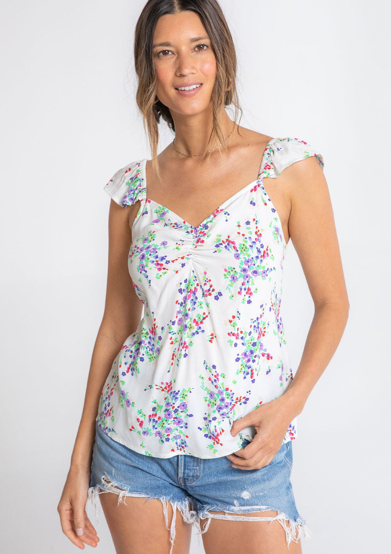 [Color: Ivory/Violet] A model wearing a white bohemian short flutter sleeve top with a purple floral print throughout and an adjustable back tie. 