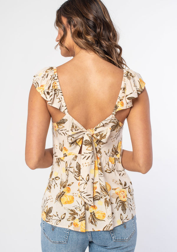 [Color: Natural/Yellow] A woman wearing a natural and yellow floral fruit print bohemian top with short flutter sleeves and a tie back detail. 